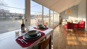 Un comedor con una mesa dispuesta para dos, ventanas amplias y un sofá rojo al fondo.