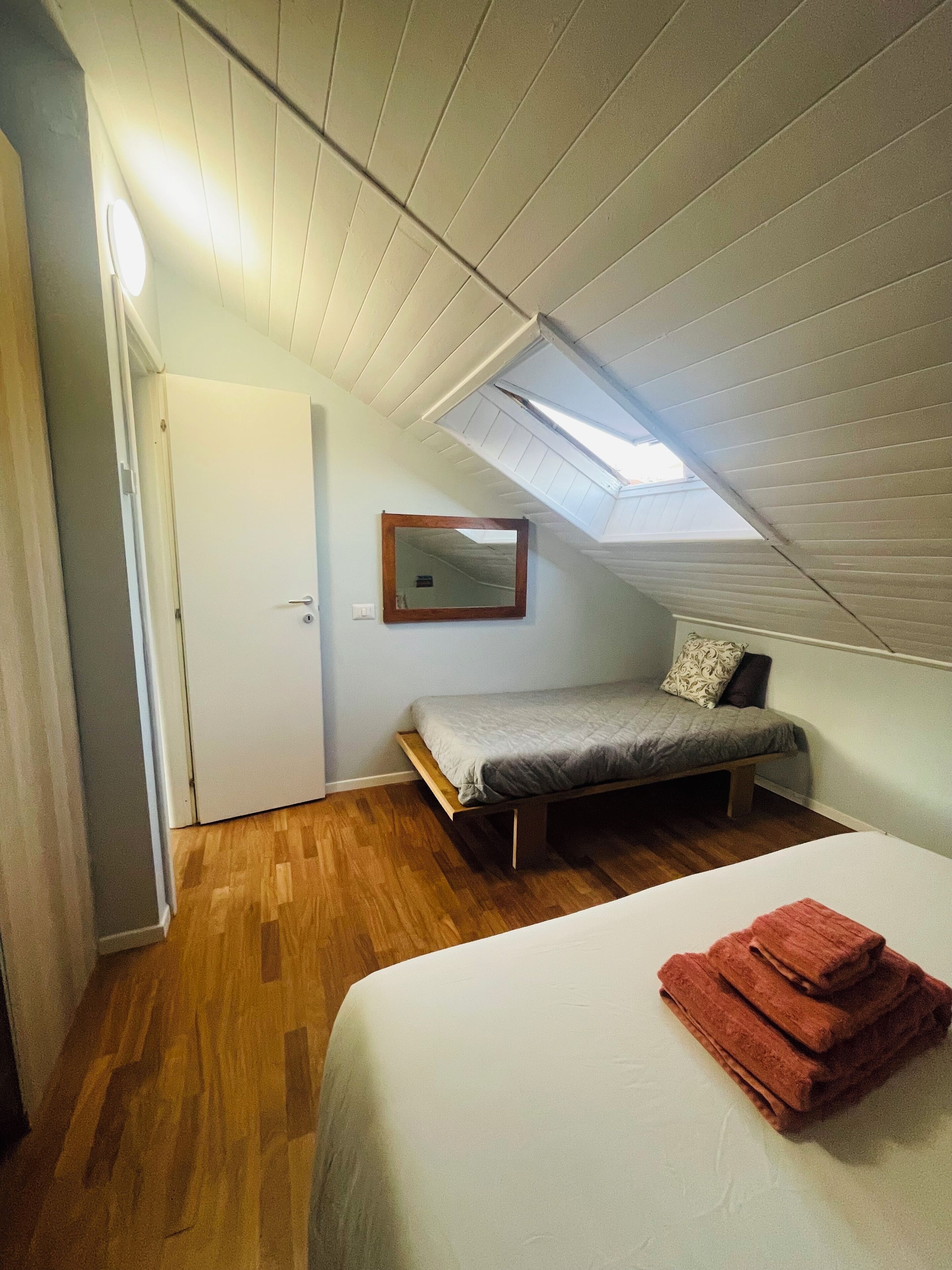 A bedroom with a sloped ceiling, a bed, a small bench, a mirror, and a skylight.