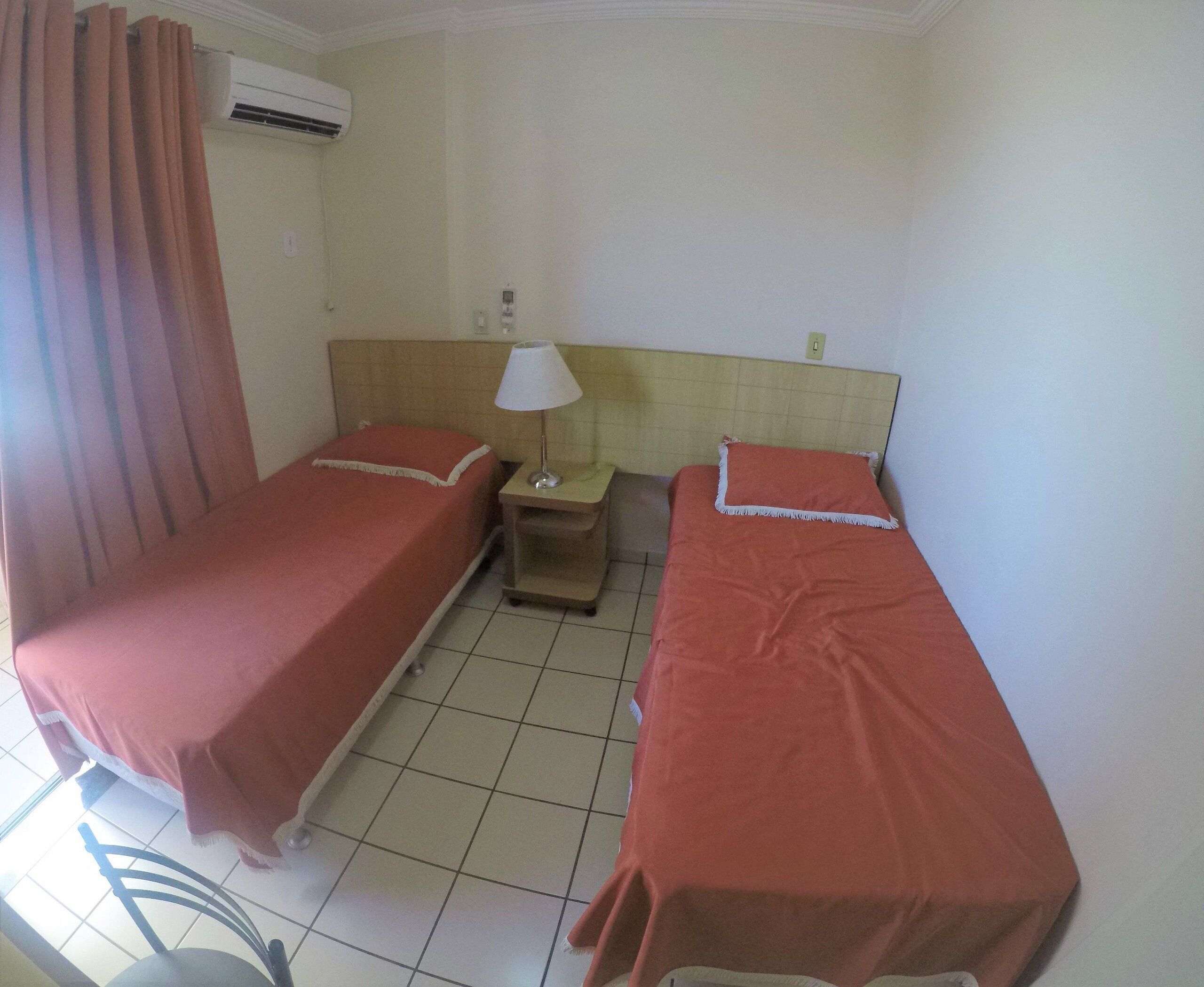 A hotel room with two single beds, a nightstand with a lamp, an air conditioning unit, and a window with curtains.