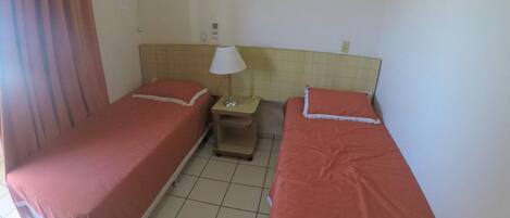 A hotel room with two single beds, a nightstand with a lamp, an air conditioning unit, and a window with curtains.