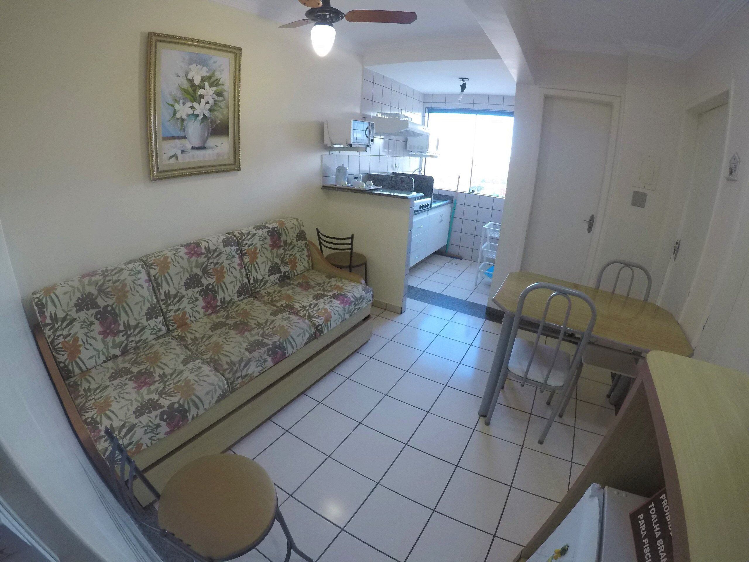 Una sala de estar con un sofá floral, una mesita redonda pequeña y un comedor con mesa de madera y sillas de metal. Se observa un ventilador de techo, y se distingue una cocina al fondo.