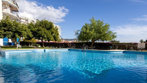 A swimming pool with clear blue water, surrounded by trees and a building with balconies.