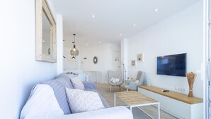 A modern living room with a sofa, chairs, a TV, and a dining area in the background.