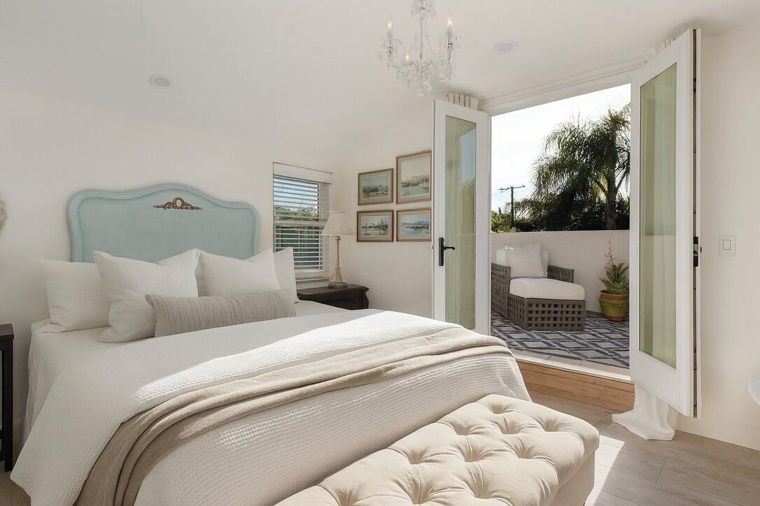 Um quarto com cama, mesa de cabeceira, um candelabro e vista para um pátio exterior através de uma porta aberta.