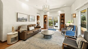 Un salon spacieux avec un canapé en cuir marron, un fauteuil bleu, une table basse ronde et un lustre. On y trouve des étagères et une vue sur l’extérieur grâce à de grandes portes-fenêtres coulissantes.