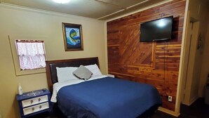 Une chambre à coucher comprenant un lit, une table de chevet, un téléviseur fixé au mur (qui est en bois) et une fenêtre avec des rideaux.
