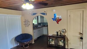 Une pièce avec un ventilateur de plafond, une chaise bleue, une table en bois agrémentée d’objets décoratifs, une porte blanche et, au fond, une cuisine.