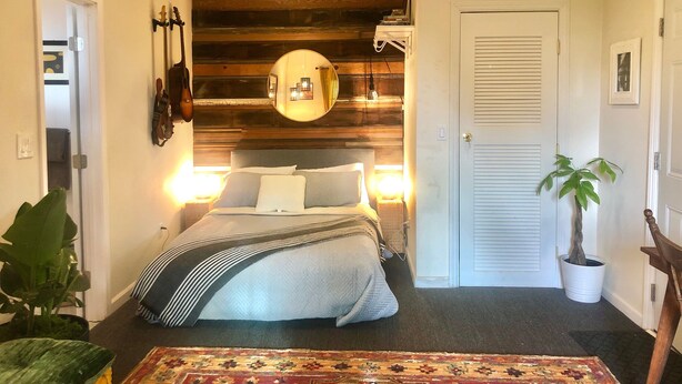 Ein Schlafzimmer mit einem Bett, Nachttischlampen, einem Spiegel, einer Zimmerpflanze und einer Tür.