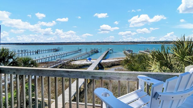 A wooden deck with white chairs overlooking a clear blue sea, a pier extending into the water, and a distant shoreline. - Waterfront 4-bedroom house on Topsail Island. Private sandbar/bring your boat! (Topsail Beach)