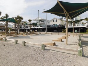 A beachfront area with wooden benches, shaded by large green canopies, and a building with a balcony in the background. - 5BR w/ 2 Decks | Porch, Bar, Printer, Walk to Beach | Site 1788 (Myrtle Beach)