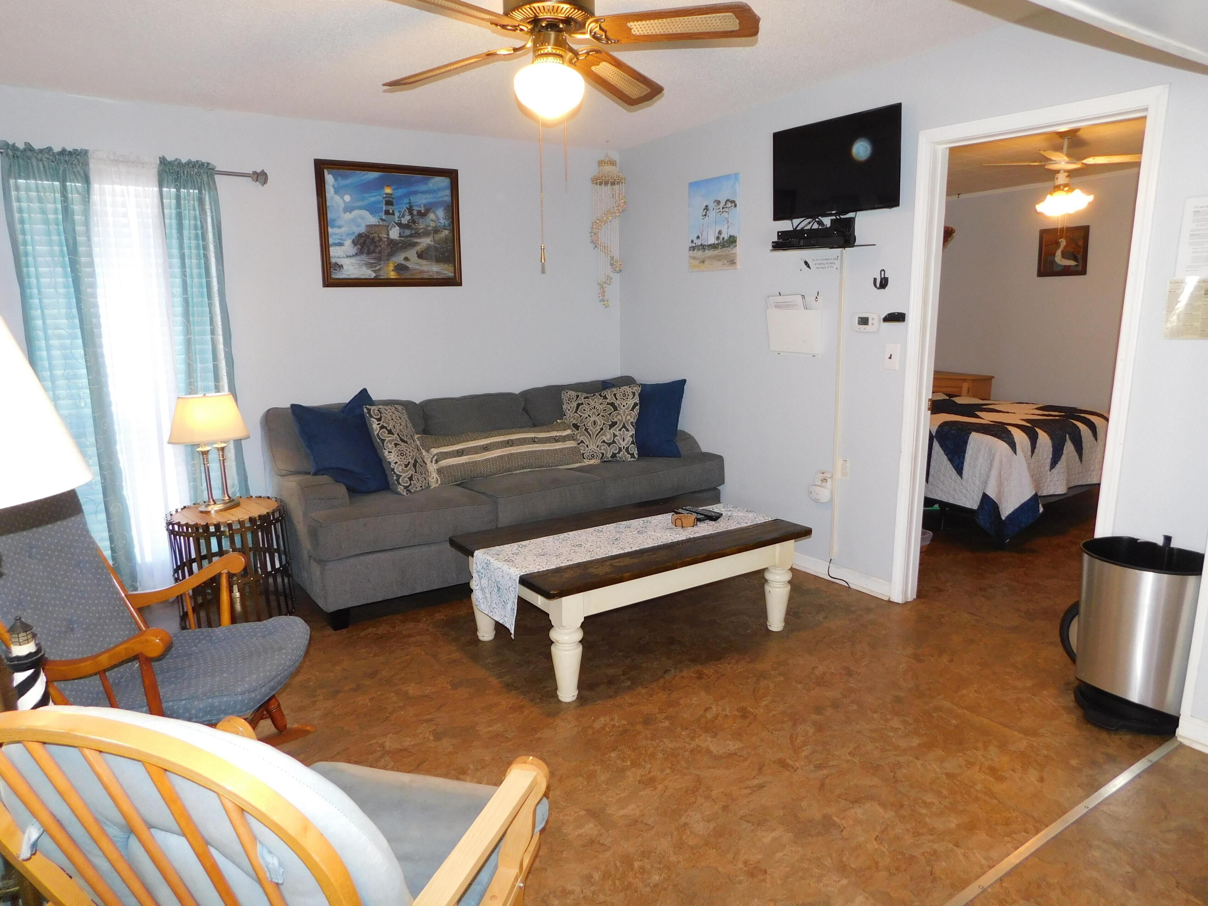 A living room with a sofa, armchair, coffee table, ceiling fan, TV, and a door leading to a bedroom.