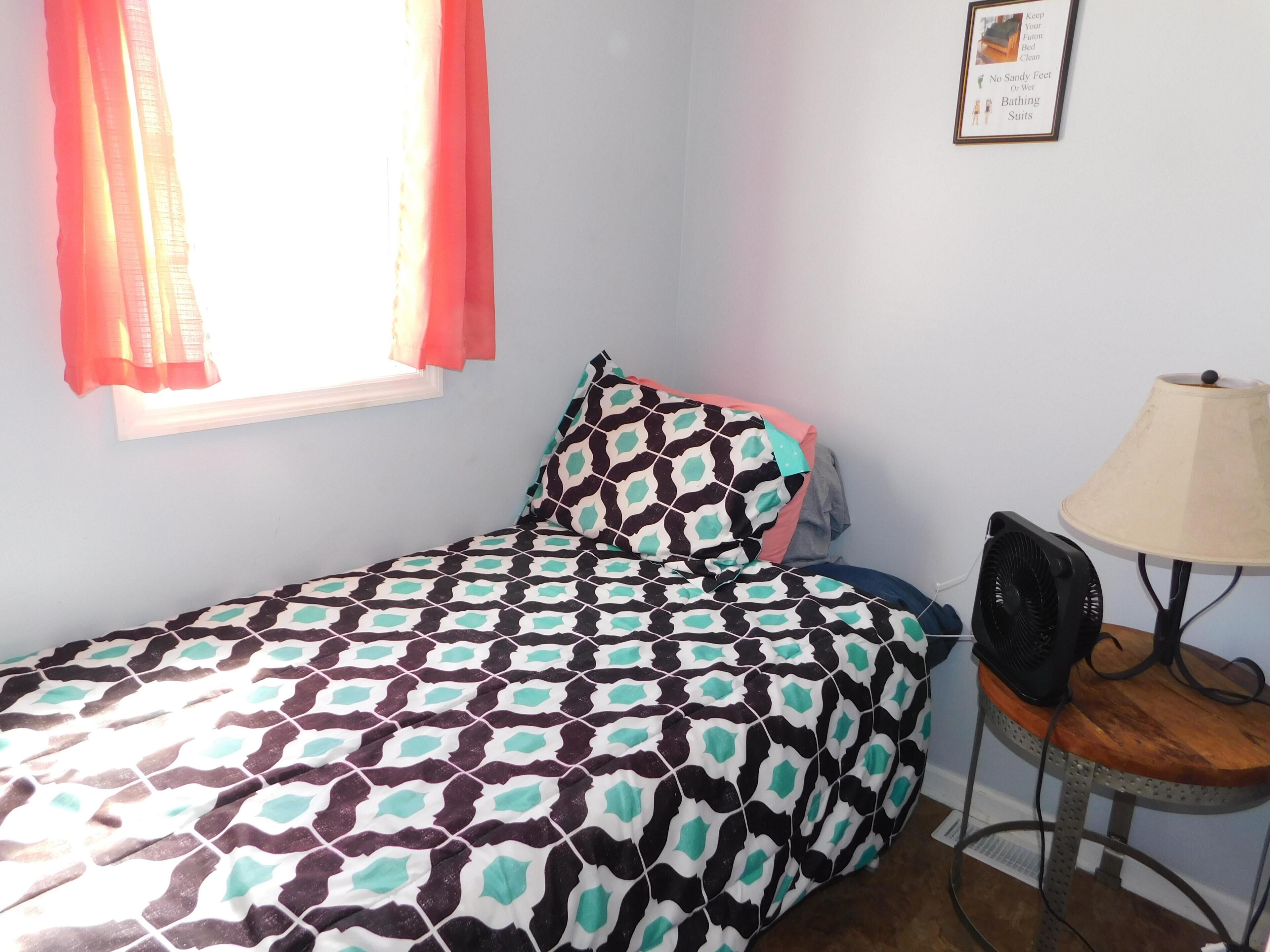 A small, neatly arranged bedroom with a single bed, a bedside table with a lamp, and a window with curtains.