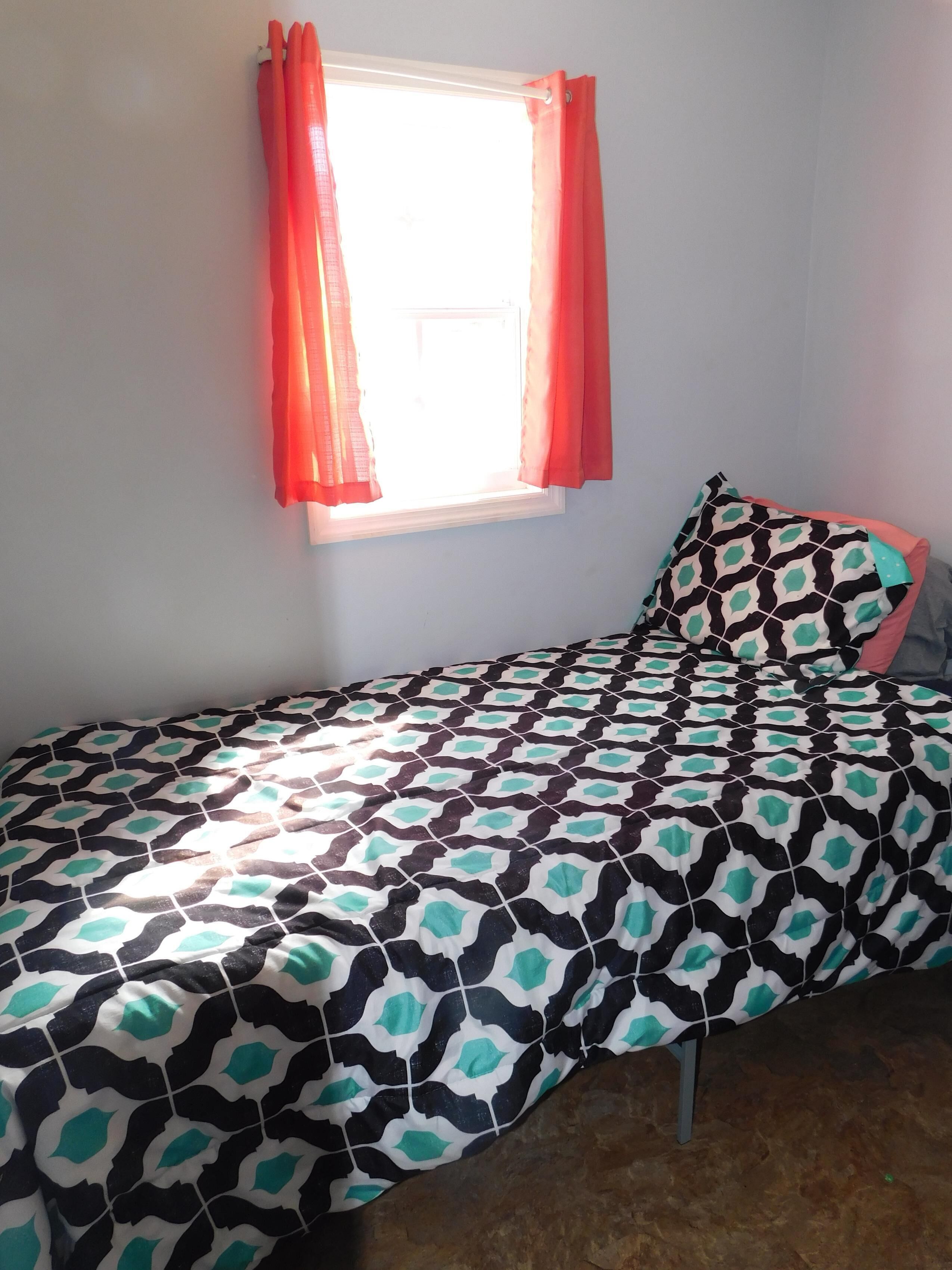 Una cama individual con una colcha estampada, una ventana con cortinas y un almohadón de diseño geométrico.