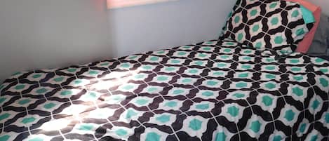 Una cama individual con una colcha estampada, una ventana con cortinas y un almohadón de diseño geométrico.