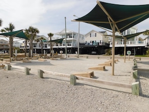 A beachfront area with wooden benches, shaded by large green canopies, and a building with a balcony in the background. - Ocean Lakes Site 1775 3bed 2bath (Myrtle Beach)