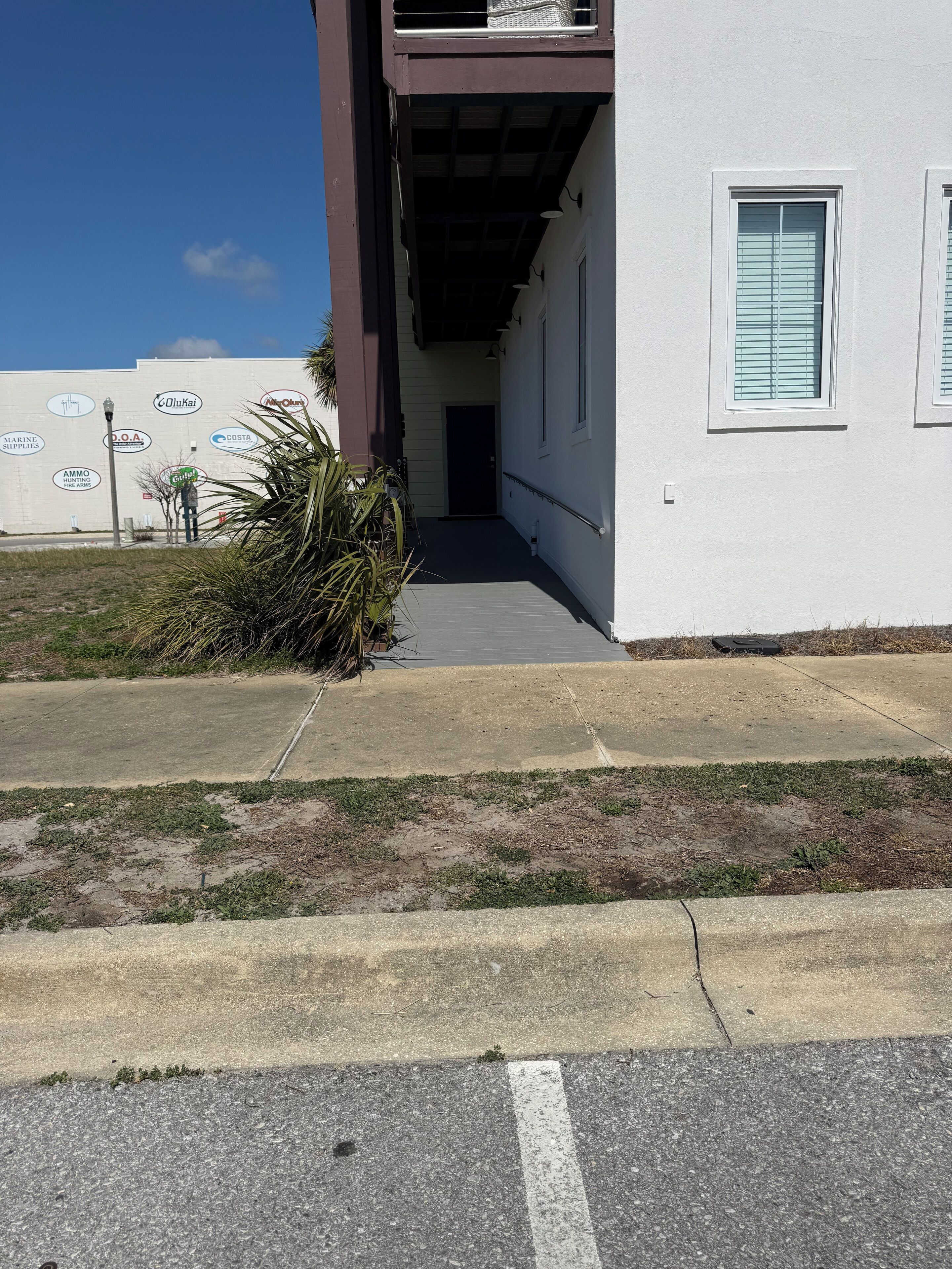 A building with a covered walkway leading to a door, a small garden area with palm plants, and a sidewalk with a curb.