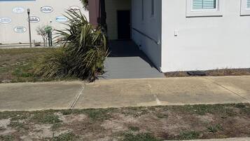 A building with a covered walkway leading to a door, a small garden area with palm plants, and a sidewalk with a curb.