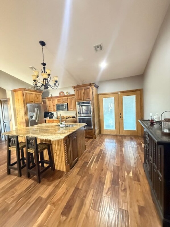 A spacious kitchen with wooden cabinets, a central island with bar stools, stainless steel appliances, and a chandelier.