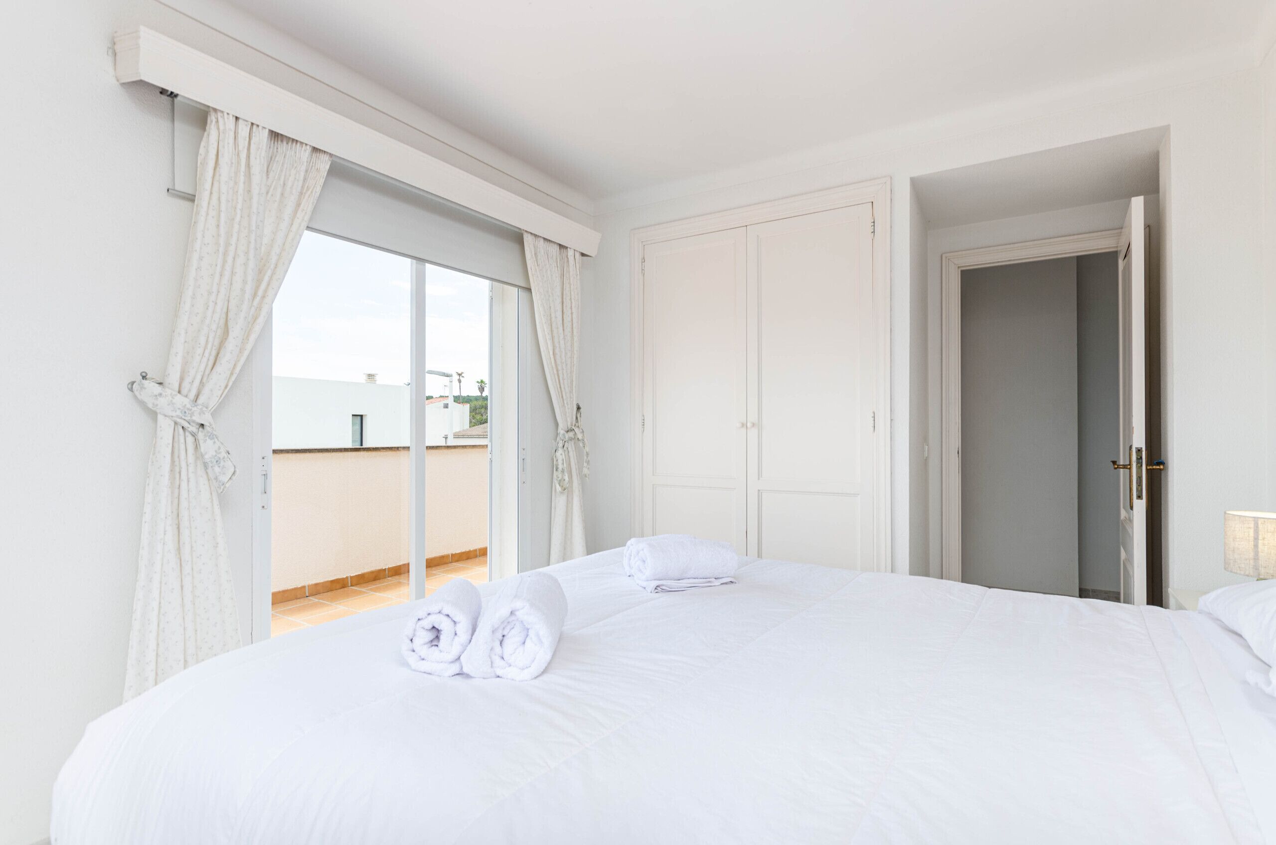 A hotel room with a large bed, white curtains, a door, and a view of the outside.