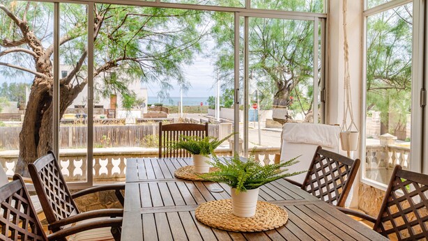 A wooden table with woven placemats and chairs, a potted plant, and a view of trees and a fence through large windows.