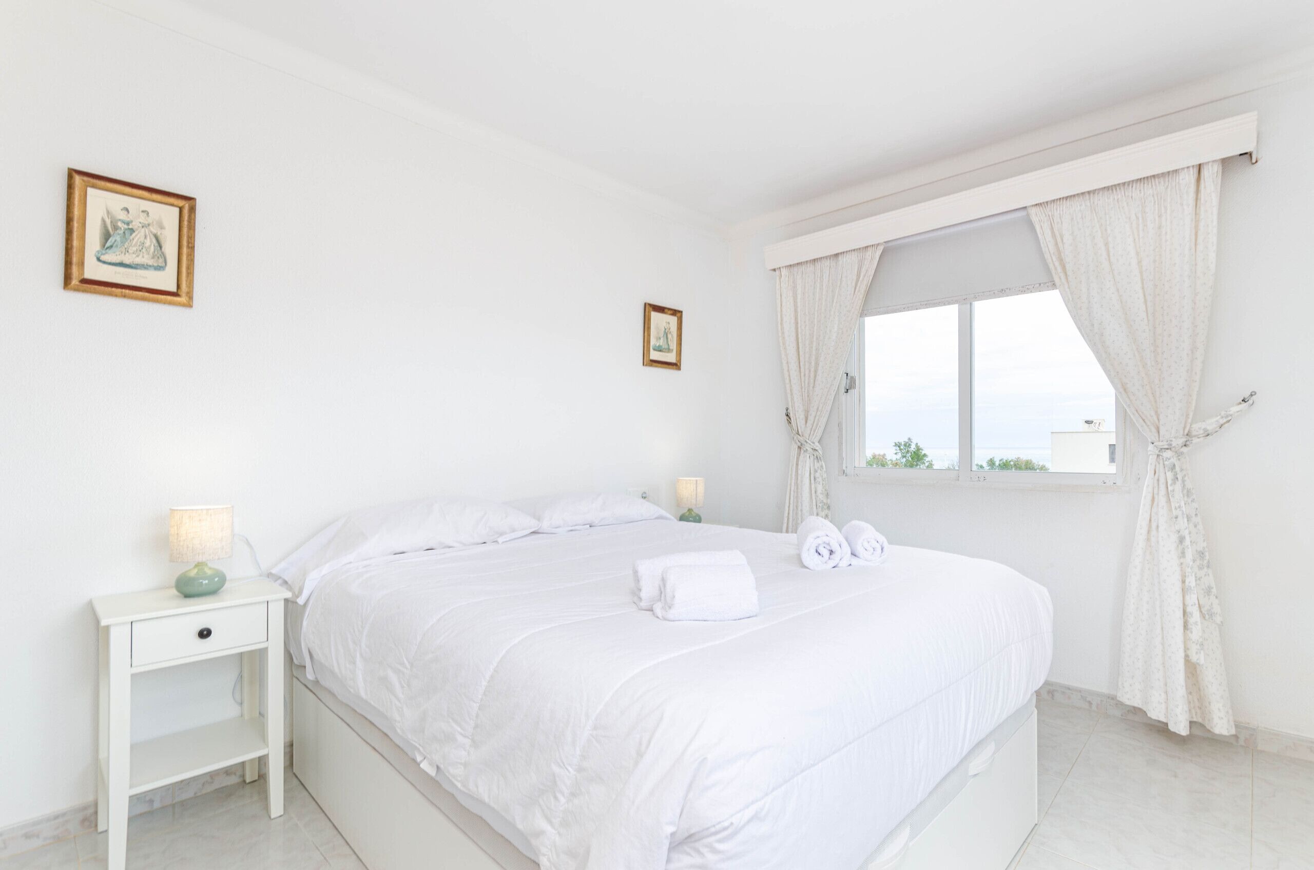 A white bedroom with a bed, a nightstand, two framed pictures, and a window with curtains.