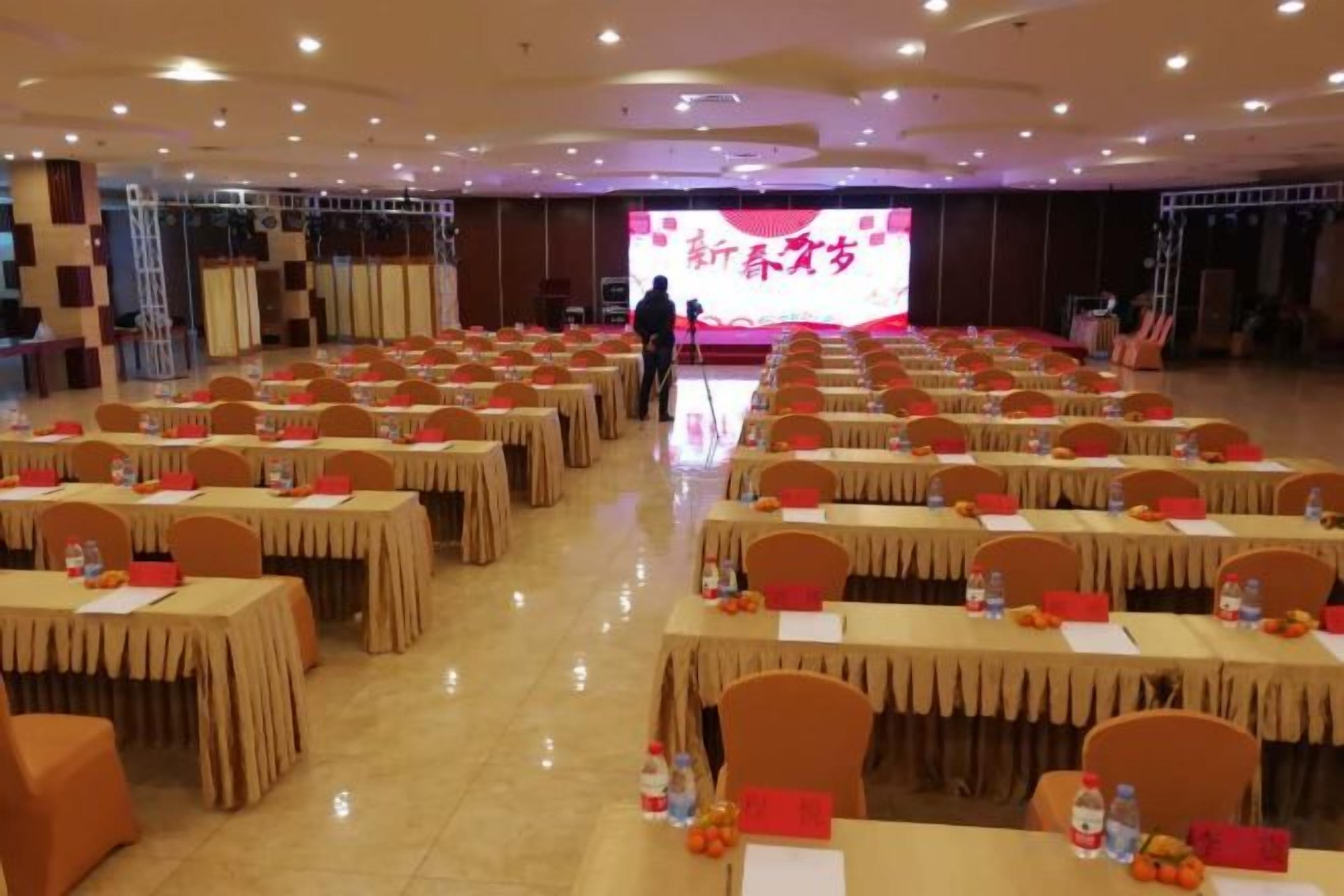 Un salón de conferencias con varias mesas y sillas, un escenario con una pantalla grande y una persona de pie cerca del escenario.