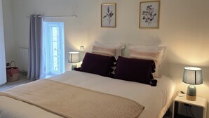Een slaapkamer met een groot bed, twee bedlampen, een nachtkastje, een raam met gordijnen en twee ingelijste botanische prints aan de muur.