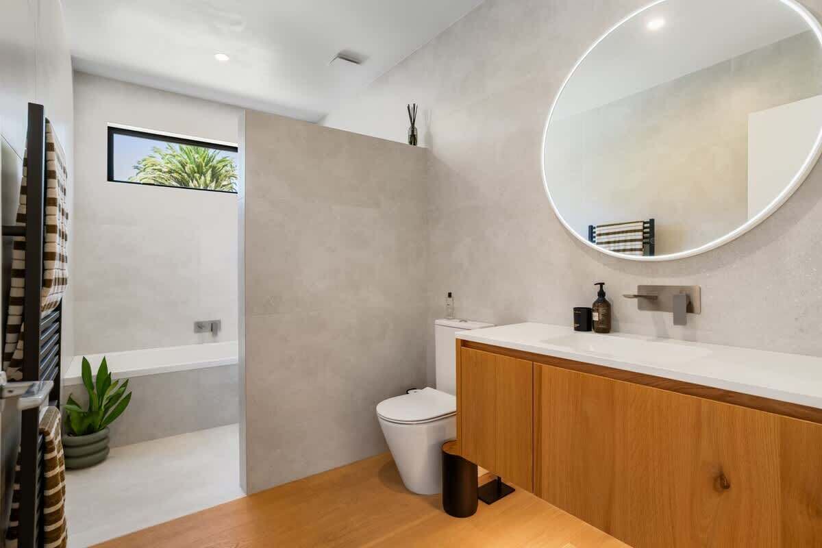A modern bathroom with a wooden vanity, a round mirror, a toilet, and a bathtub.
