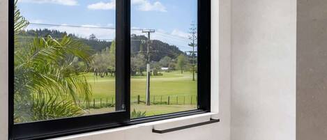 A room with a large window, a green object on the floor, and a view of a green field and trees.