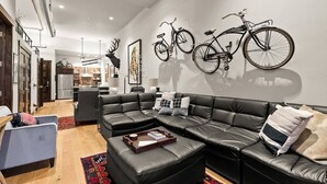 A modern living room with a sectional sofa, a coffee table, and a dining area in the background. - Downtown Barnwood Condo. WSTR-21-00029 (Whitefish)