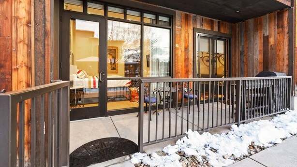 A wooden exterior with a glass door entrance, a covered patio with metal railings, and a snow-covered ground. - Downtown Barnwood Condo. WSTR-21-00029 (Whitefish)