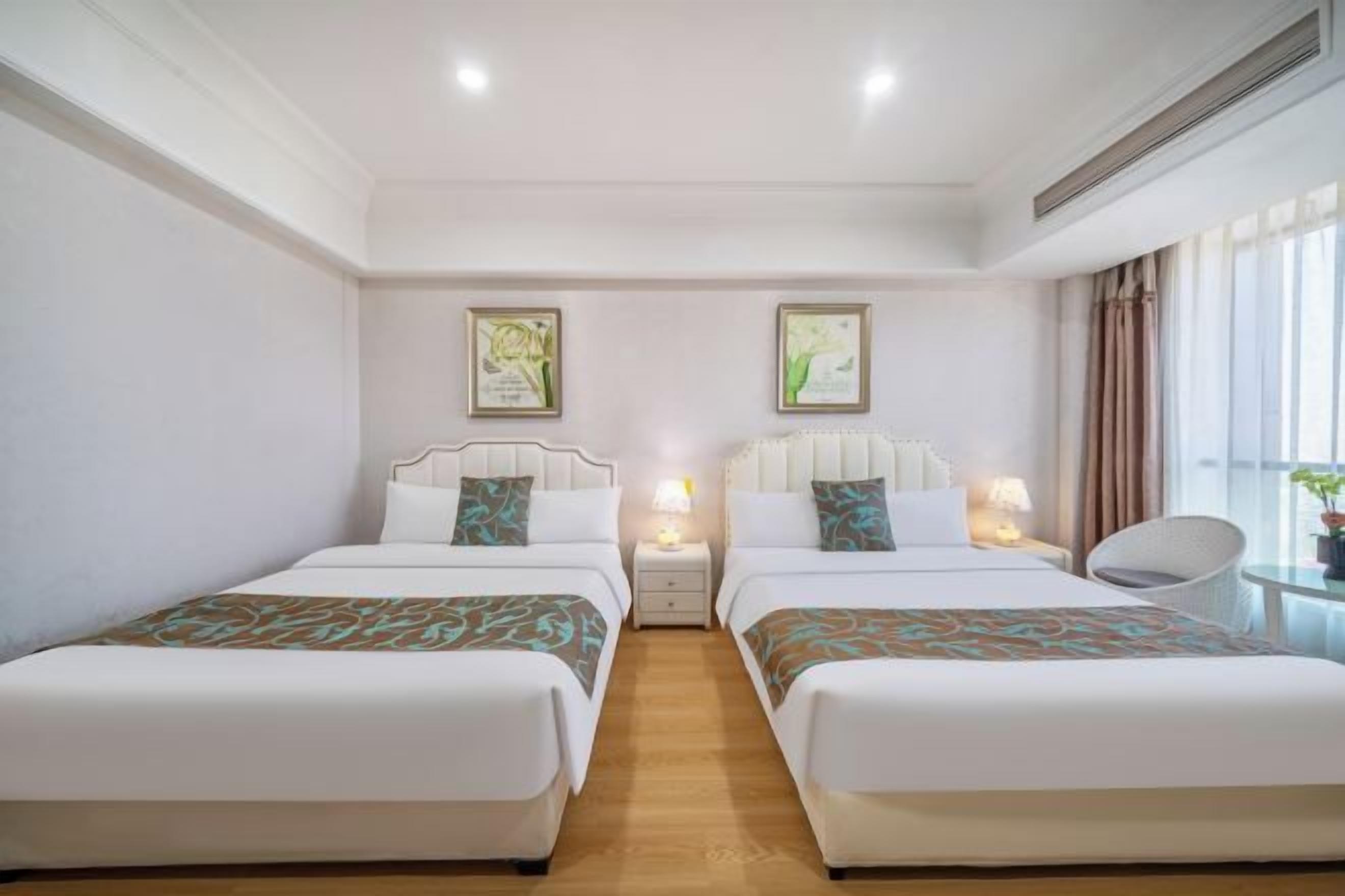 Habitación de hotel con dos camas, una mesita de noche con lámpara, una silla y dos cuadros en la pared.