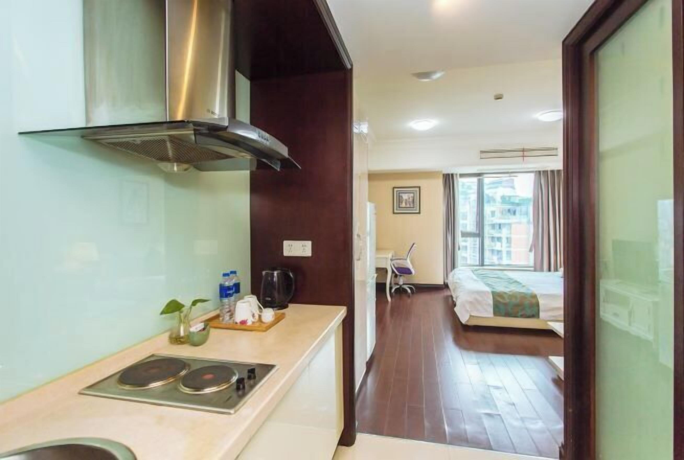 A compact hotel room with a kitchenette, a bed, a chair, and a view of the cityscape.