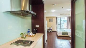 A compact hotel room with a kitchenette, a bed, a chair, and a view of the cityscape.