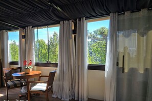 Una habitación con una mesa redonda, dos sillas, una planta en maceta y vistas a árboles a través de ventanas amplias con cortinas translúcidas.
