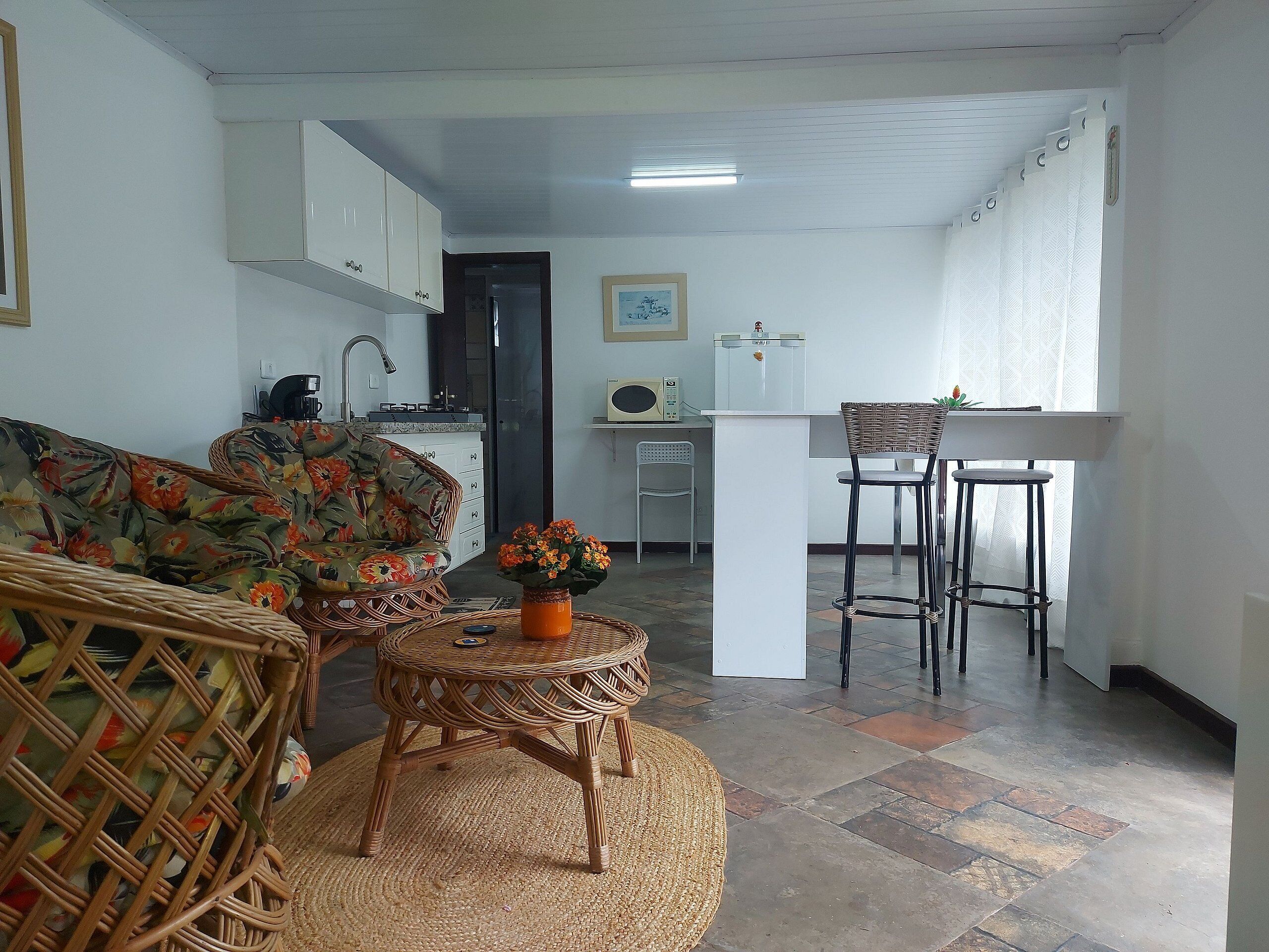 Una sala de estar con un sofá, una silla de mimbre, una pequeña mesa redonda con un jarrón de flores, y una cocina con un refrigerador, estufa y un taburete de bar.