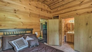 A wooden cabin bedroom with a bed, pillows, a nightstand, and a door leading to a bathroom with a sink and mirror. - Cozy cabin 1 bedroom, WiFi, AC above Sylamore Creek in welcoming Mountain View (Mountain View)
