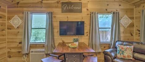 A wooden cabin interior with a dining table, two chairs, a sofa, and a TV.