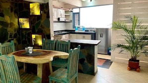 Een eetgedeelte met een houten tafel en stoelen, een keuken op de achtergrond, en een potplant.