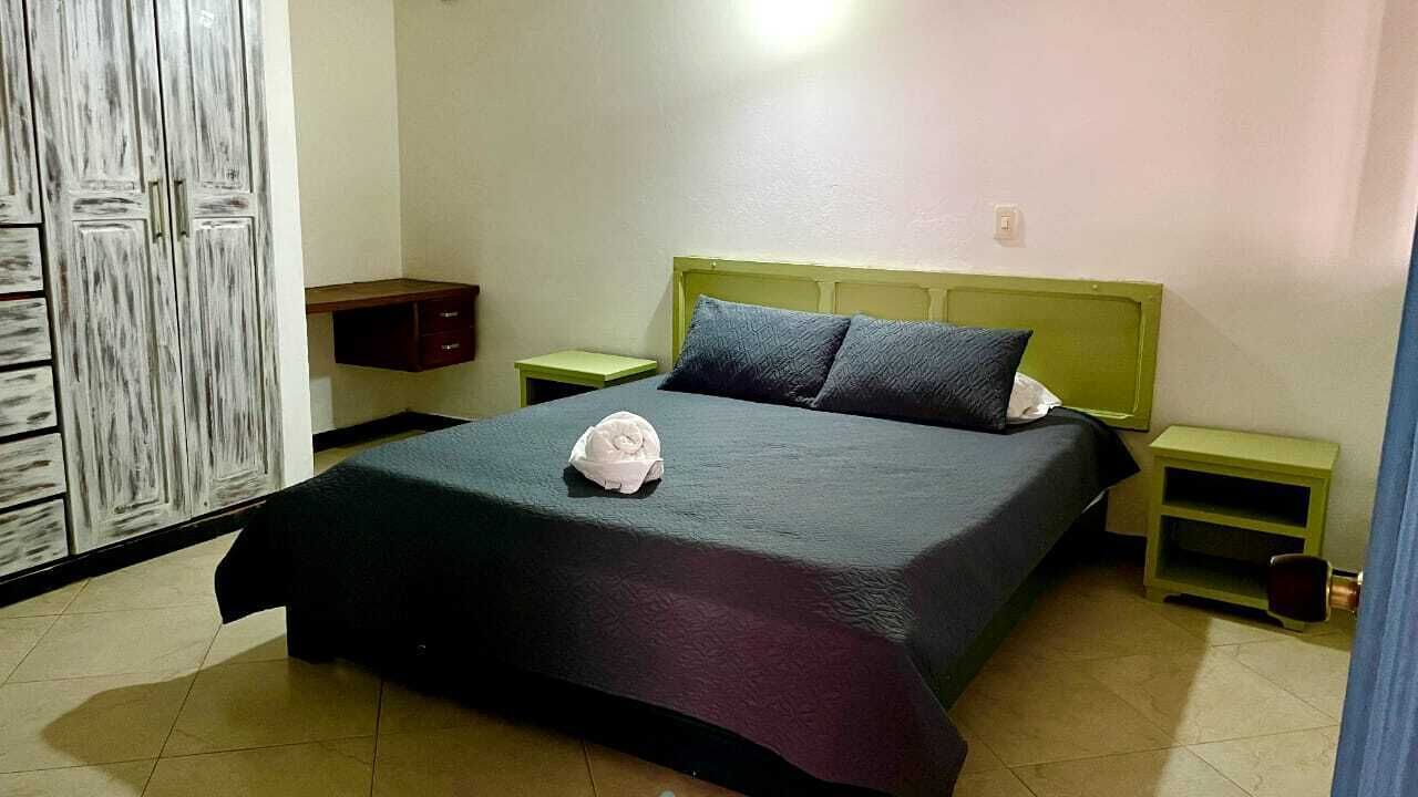 Una cama bien hecha con una colcha oscura y dos almohadas, un armario de madera y una mesita de noche con una lámpara.