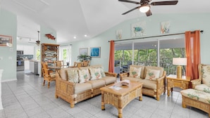 A spacious living room with a large sofa, two armchairs, a coffee table, and a dining table. There is a ceiling fan, a kitchen in the background, and a large window with curtains. - Tropical paradise on canal in Lower Keys-pool, tiki, firepit, dock, dog friendly (Summerland Key)