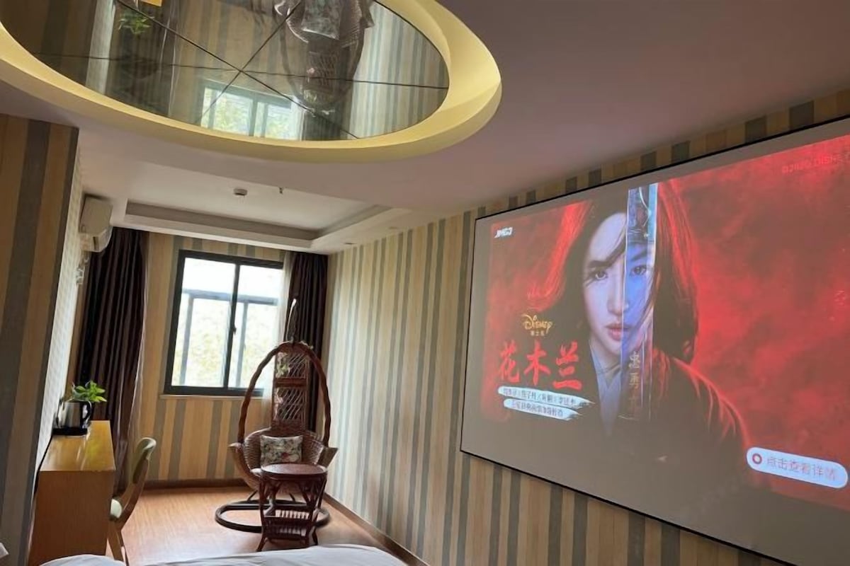 A hotel room with a large screen displaying a movie, a wooden chair, a desk with a lamp, and a window with curtains.