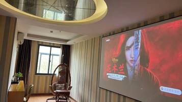 A hotel room with a large screen displaying a movie, a wooden chair, a desk with a lamp, and a window with curtains.