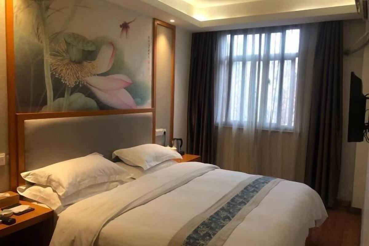 Hotellihuone, jossa on sänky, yöpöytä, taulu, ikkuna verhoilla ja seinälle asennettu televisio.