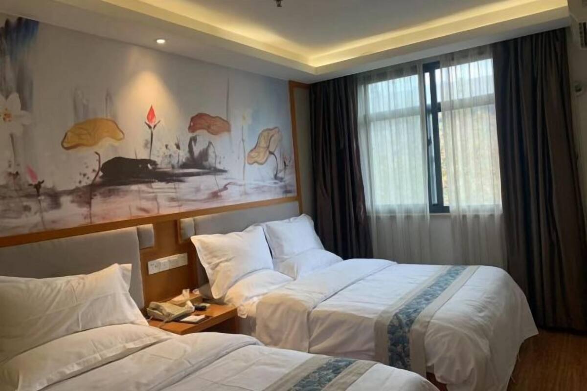 Hotellihuone, jossa on kaksi sänkyä, yöpöytä, seinämaalaus ja suuri ikkuna, jossa on verhot.