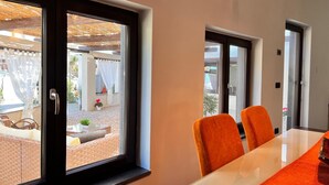Un espace repas moderne avec une table en verre et des chaises orange, de grandes fenêtres avec des rideaux et une vue sur un patio extérieur.