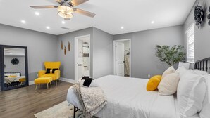 Une chambre à coucher avec un lit, un ventilateur de plafond, un miroir, un fauteuil jaune et une plante en pot.