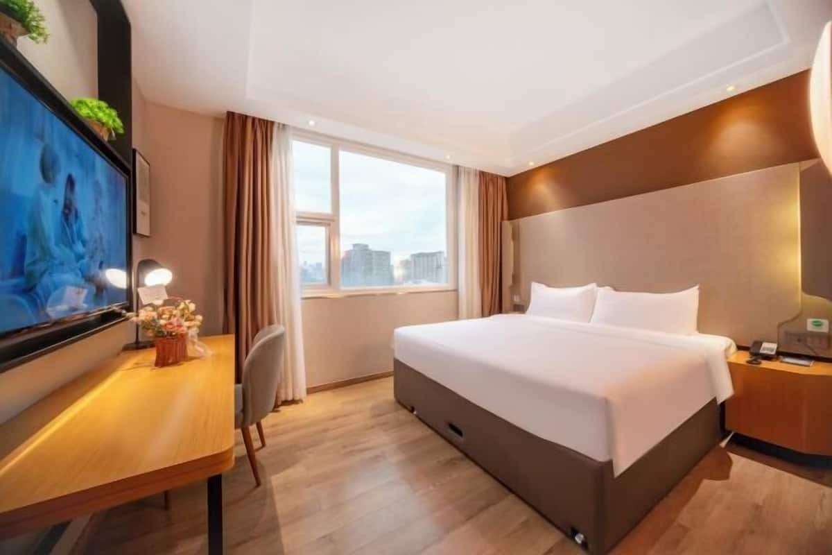 A hotel room with a bed, a desk, a TV, and a window with a city view.
