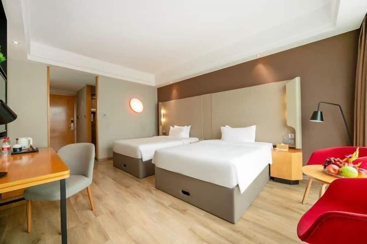 A hotel room with two beds, a desk, a chair, a small table with fruits, and a mirror.