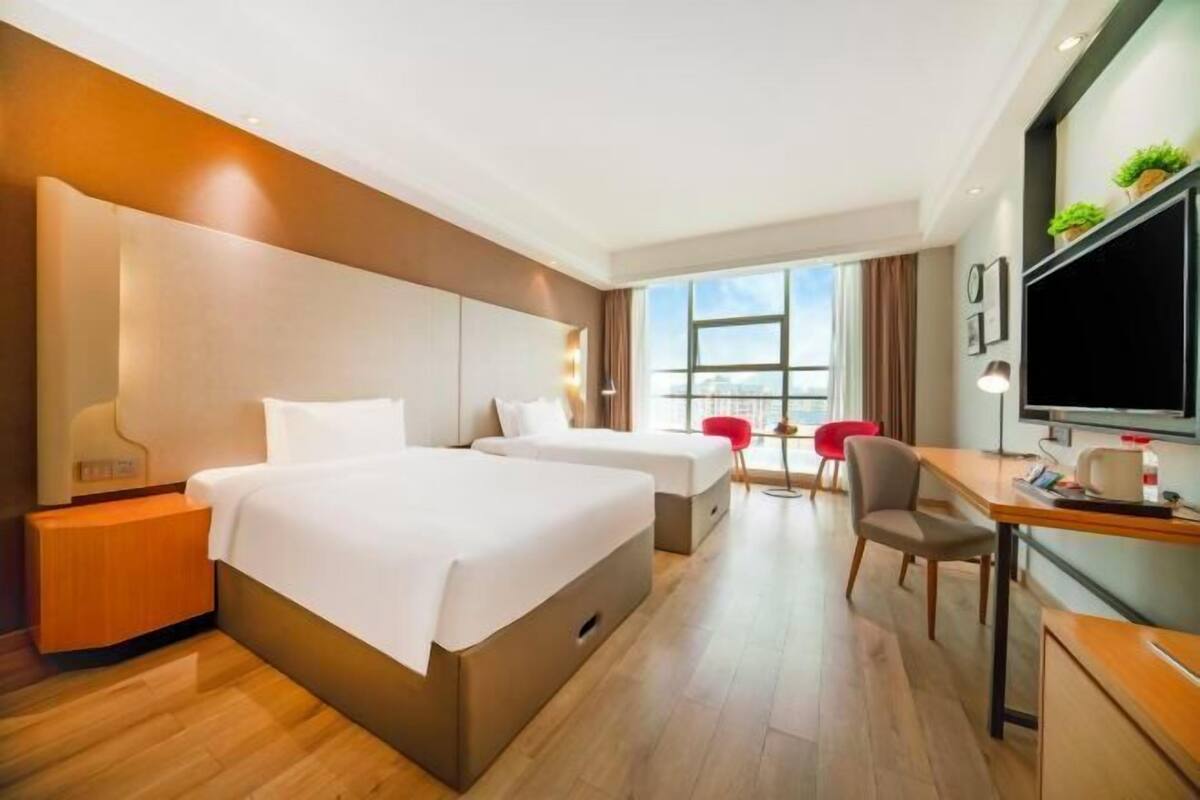 A hotel room with two beds, a desk, a chair, a TV, and a window with a city view.